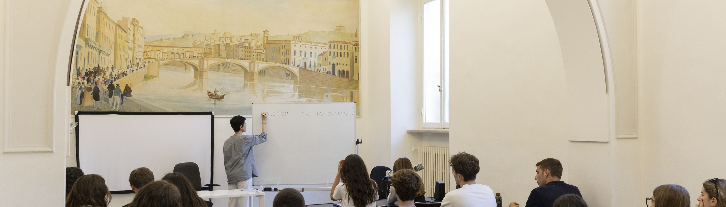A lesson in the frescoed classroom of Unicollege, with the painted Ponte Vecchio behind the lecturer